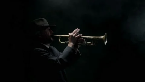 Jazzman in jacket and hat plays trumpet on dark background in the fog, side view Stock Footage 163286379