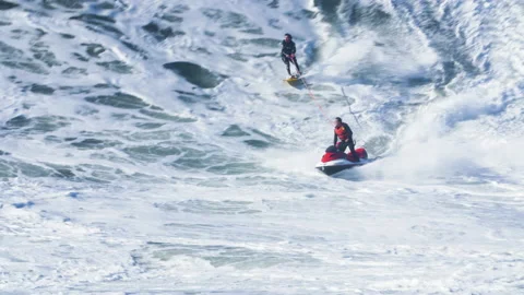 Jet ski driver riding on huge ocean wave with pro surfer wakesurfing behind him Stock Footage 155662823