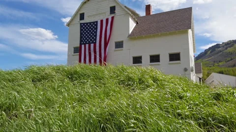 Jib up from tall grass to reveal rustic barn displaying huge American flag Stock Footage 202152032