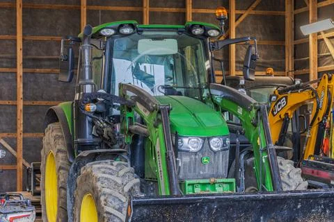 John Deere tractor with front loader parked beside JCB excavator inside barn. Stock Photos