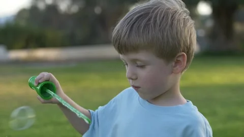 A joyful 4-year-old boy blows soap bubbles in a sunny park, surrounded by green Stock Footage 317319343