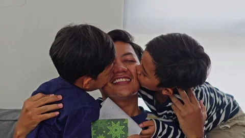 Joyful Asian dad receiving present and hugs from children at home on sofa 库存影片 335179418