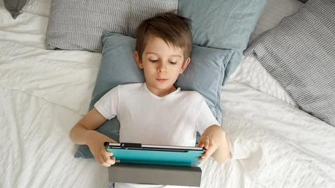 A joyful boy is happy using a tablet computer while lying on his bed during.. Stock Photos