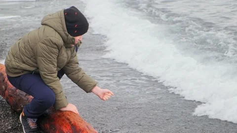 Joyful boy in hat and jacket plays with sea water waves 库存影片 134268295