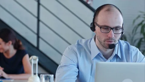 Joyful call centre agent with his headset talking looking at laptop Stock Footage