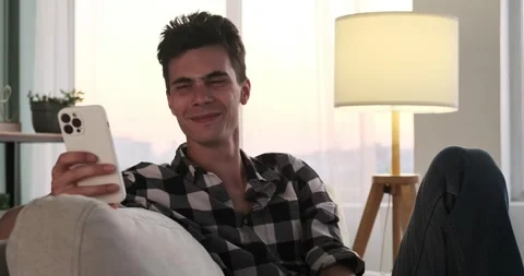 Joyful Caucasian Man Laughing While Using Phone on Sofa Stockbeeldmateriaal 250026799