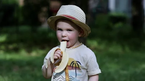 A joyful child in a hat eats a banana and smiles Stock Footage 157236994