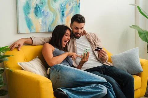 Joyful couple on a yellow sofa using phones together while smiling at a shared Stock Photos