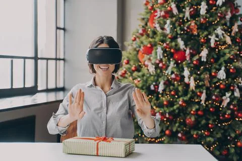 Joyful hispanic female in VR headset sitting at desk with Xmas present during Foto stock