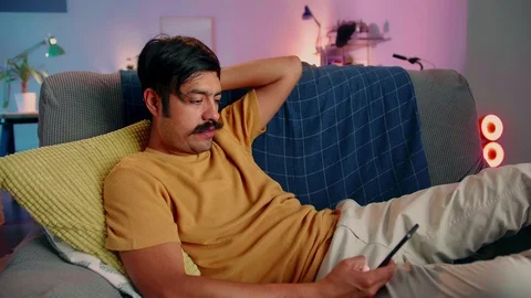Joyful Hispanic guy chilling on sofa with black mobile phone in hands at home Stock Footage 117131235