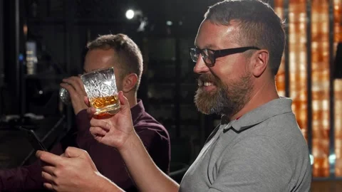 Joyful man savoring whiskey while texting on his smartphone Stock Footage 311674502