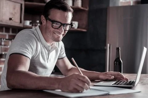 Joyful millennial guy taking notes while working on laptop Stock Photos