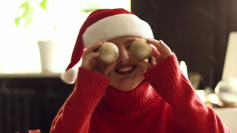 Joyful person in Santa hat playfully covers eyes with Christmas ornaments Stock Footage 339079088