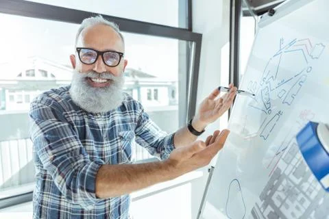 Joyful pleasant elderly worker is standing near flip chart Stock Photos