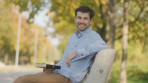 Joyful reader on a park bench, showing off his book against a fall backdrop Stock Footage 300252276