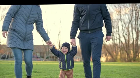 A joyful toddler walks between two adults, holding their hands in a grassy park Vidéo 279972921