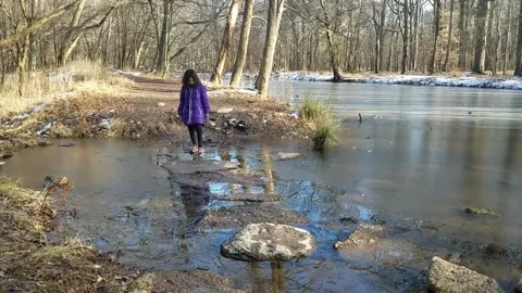 Joyful toss of thin ice sheet by young girl balanced on flat stone in lake Stock Footage 201644731