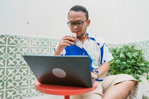 Joyful young Asian man in a patterned shirt is using his laptop to work whi.. Stock Photos