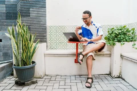 Joyful young Asian man in a patterned shirt is using his laptop to work whi.. Stock Photos