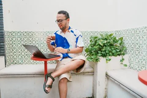 Joyful young Asian man in a patterned shirt is using his laptop to work whi.. Stock Photos