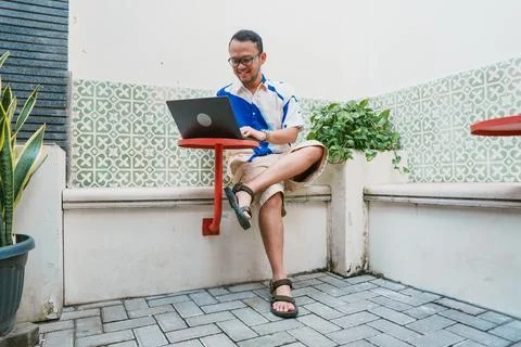 Joyful young Asian man in a patterned shirt is using his laptop to work whi.. Stock Photos