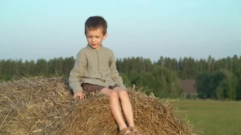 Joyful young boy sits on a large hay bale in a sunny rural field Video stock 342966474