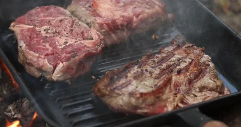 Juicy beef steaks sizzling on a hot cast iron grill pan with flames Stock Footage 339074219