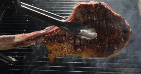 Juicy tomahawk steak grilling on barbecue grid. Chef hand flips roasting steak Stock Footage 275657058