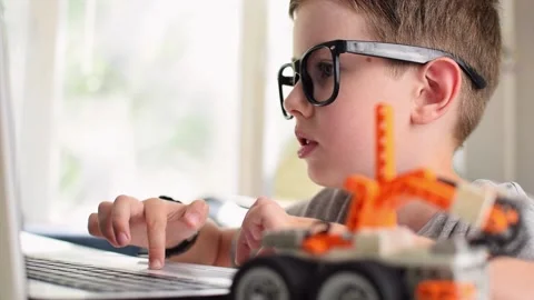 Keen boy in glasses programs a robot car and typing at laptop at home. 스톡 동영상 196793025