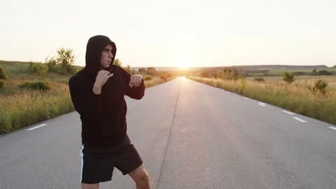 Kickboxer with hood on head training kicks on country road singly during sunset Stock Footage 139799498