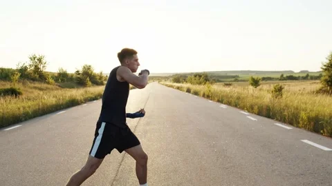 Kickboxer with wrapped hands in tapes training kicks on country road singly Stock Footage 139956045