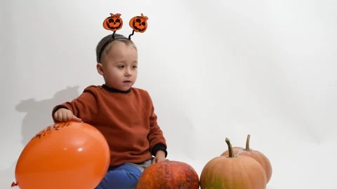 Kid boy of 3 years  sits at halloween pumpkins and catches balloon. Halloween Stock Footage 253990735