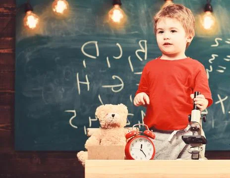Kid boy near microscope in classroom, chalkboard on background. First former Stock Photos