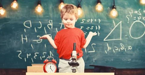 Kid boy near microscope, clock in classroom, chalkboard on background. First Stock Photos