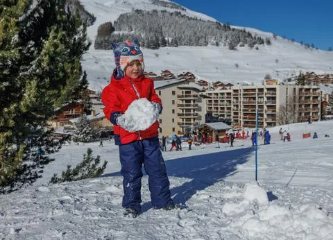 Kid boy in red jacket holding a giant snowball on sunny slope Stock Photos