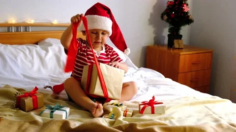 A kid in a Christmas hat sits on the bed and unpacks the gift. Stock Footage 163831877