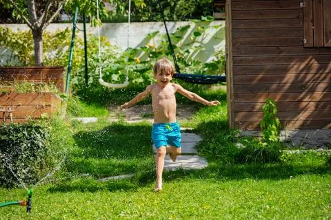 Kid enjoy sprinkle of water while sprinting across a green lawn Stock Photos