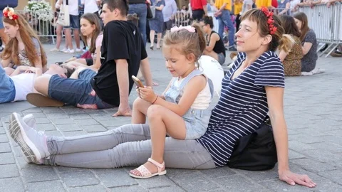 Kid girl enjoy play phone game her mother sit on cobblestone listen to concert Stock Footage