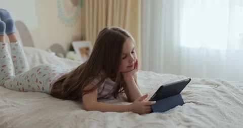 Kid girl using digital tablet watching cartoons lying on bed at home. Stock Footage 335644073