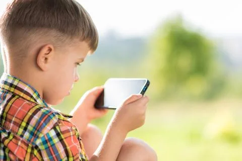 The kid looks into the mobile while sitting in the summer on the street. Smal Stock Photos