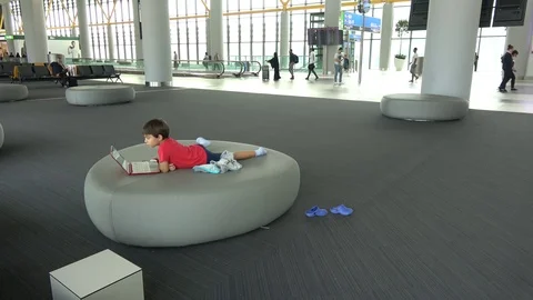 Kid with notebook rests on round sofa during layover in departure area Stock Footage 115658038