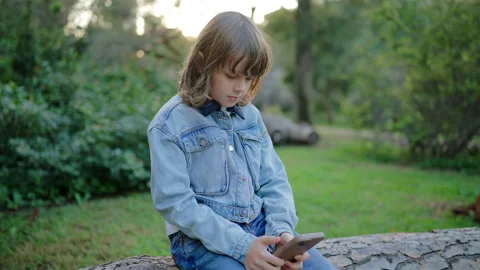 Kid on the phone sitting on fallen tree log in green park at sunset. Vídeo Stock 338140502