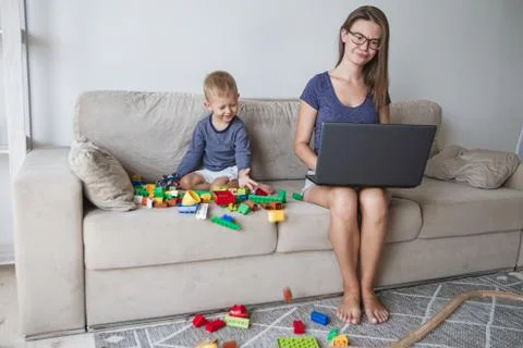 Kid plays on sofa while mom works with laptop Stock Photos