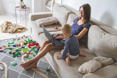 Kid plays on the sofa while mom works with laptop, distracts and Stock Photos