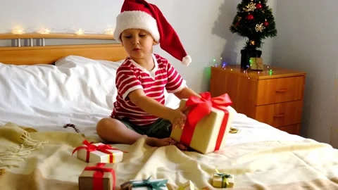 A kid, sitting on the bed, looking at and shaking boxes of Christmas gifts Video stock 163831833
