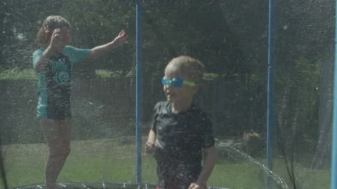 Kids bouncing on trampoline playing in summer with water hose swimming Stock Footage 135789159