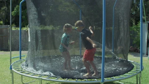 Kids bouncing on trampoline playing in summer with water wide shot Stock Footage 135791231