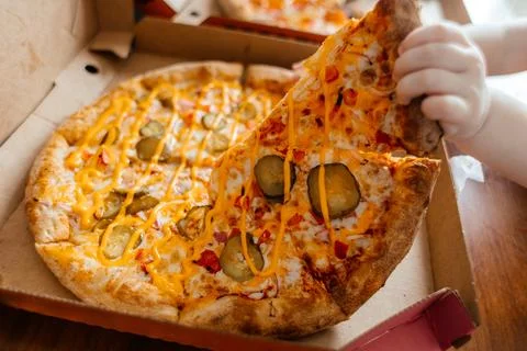 A kids hand pulls a slice of pizza from a box. The pizza has cheese, pickles, Stock Photos