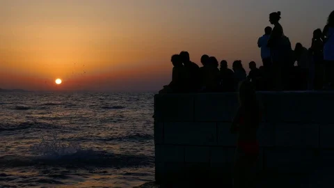 Kids jumping in water while crowd of people sitting together looking at sunset Stock Footage 89160393