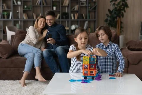 Kids play constructor while parents use smartphone seated on sofa Stock Photos